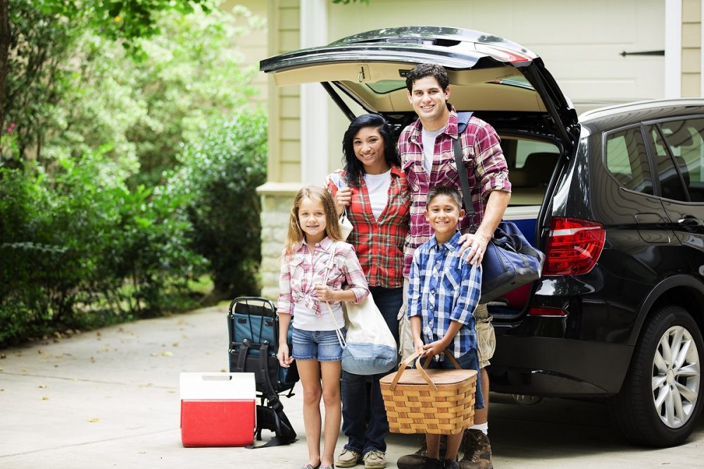 family of two adults and two children standing by car trunk with luggage, a cooler, and a picnic basket