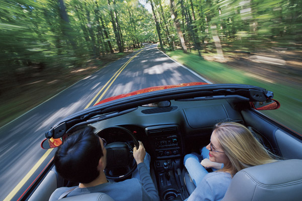 Two people driving along a wooded road in a convertible