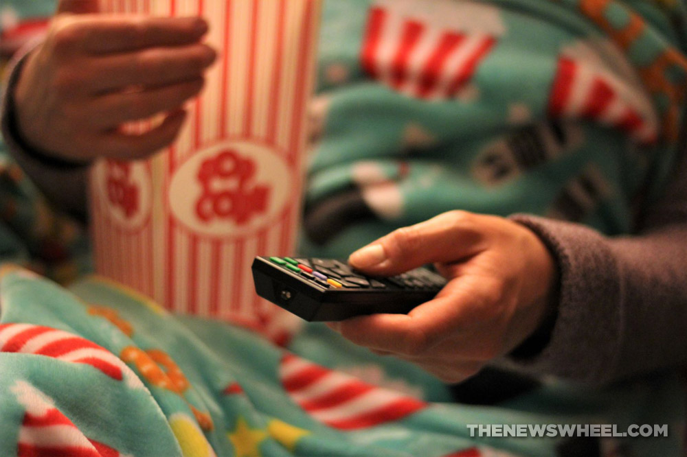 Person with remote control, a container of popcorn, and a blanket sitting on a chair or couch