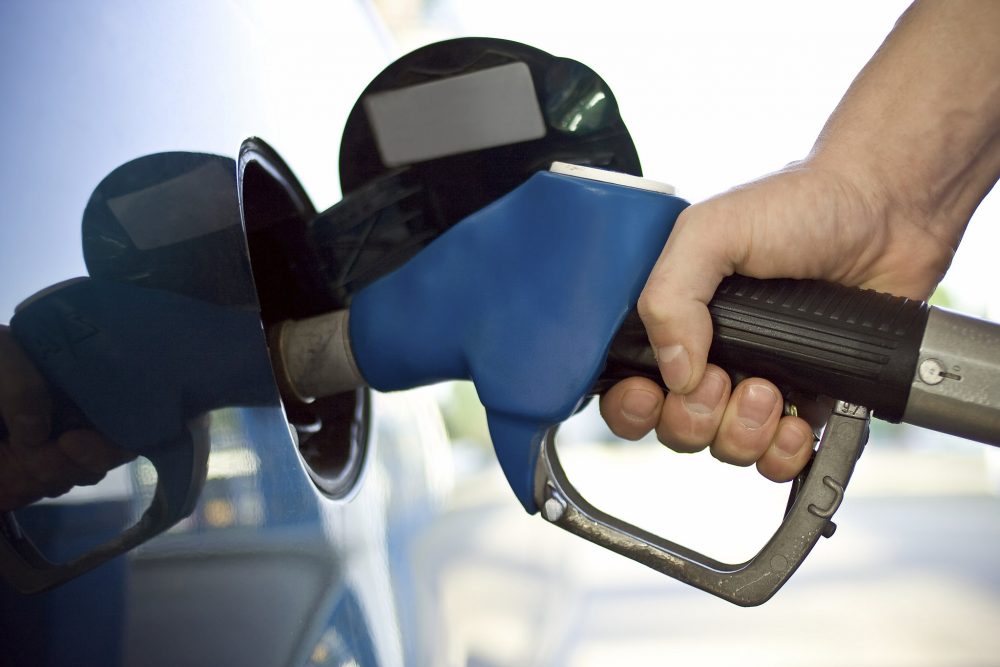 A person's hand grips the handle of a blue fuel dispenser with the nozzle inserted in the vehicle fuel filler 