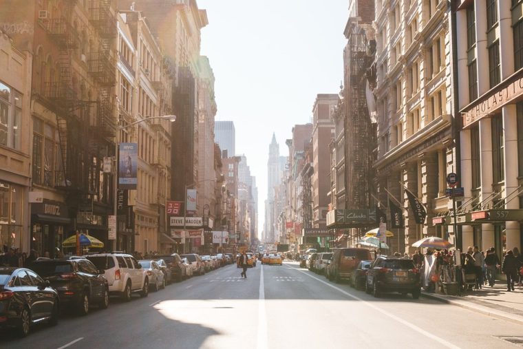 View of a city street with cars parked down the side and tall buildings on each side.