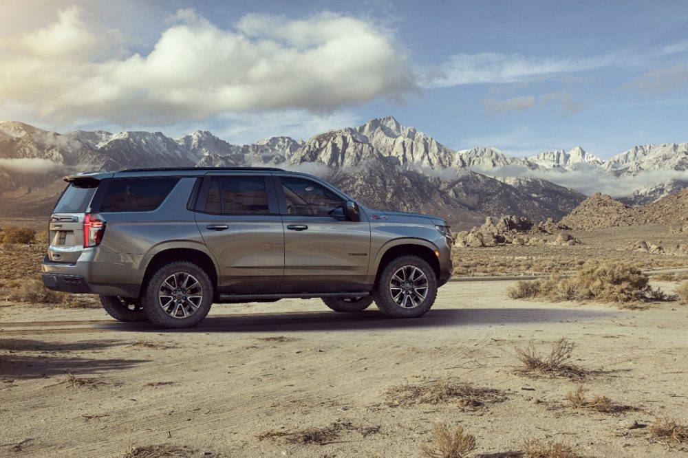 Sideview of a silvery grey 2021 Chevrolet Tahoe Z71 SUV in a desert landscape with mountains in the distance under a partly cloudy blue sky