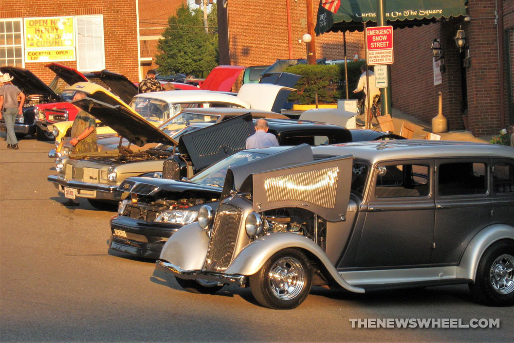 Classic cars show on city streets with hoods up and spectators walking