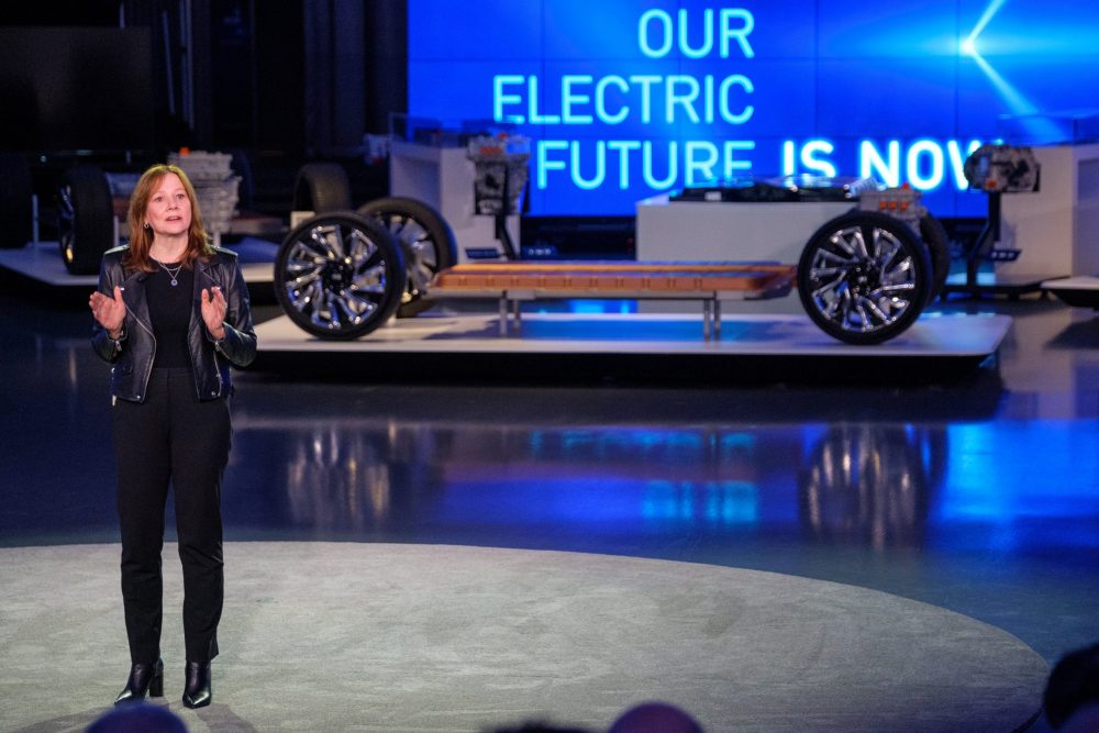 Mary Barra speaking in front of a car platform and a sign saying, "Our Electric Future Is Now"