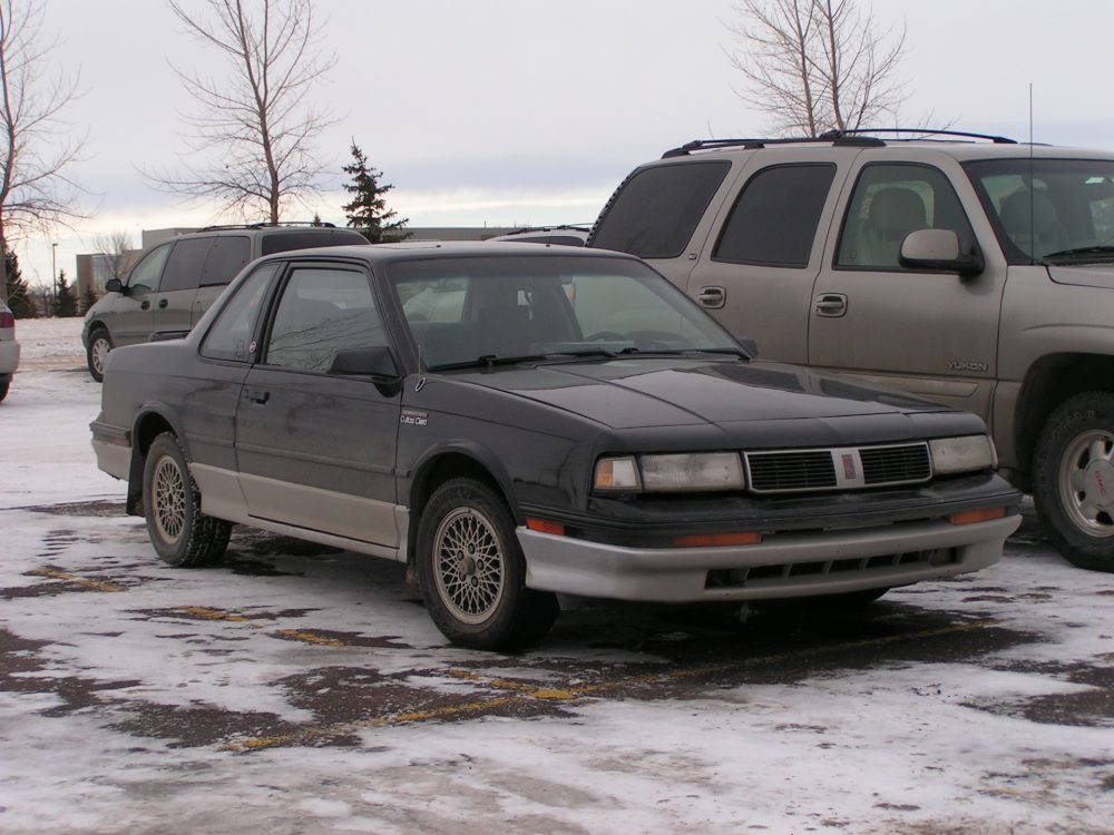 a 1987 Oldsmobile Cutlass in a snowing parking lot, reminiscent of a scene from 'Fargo"