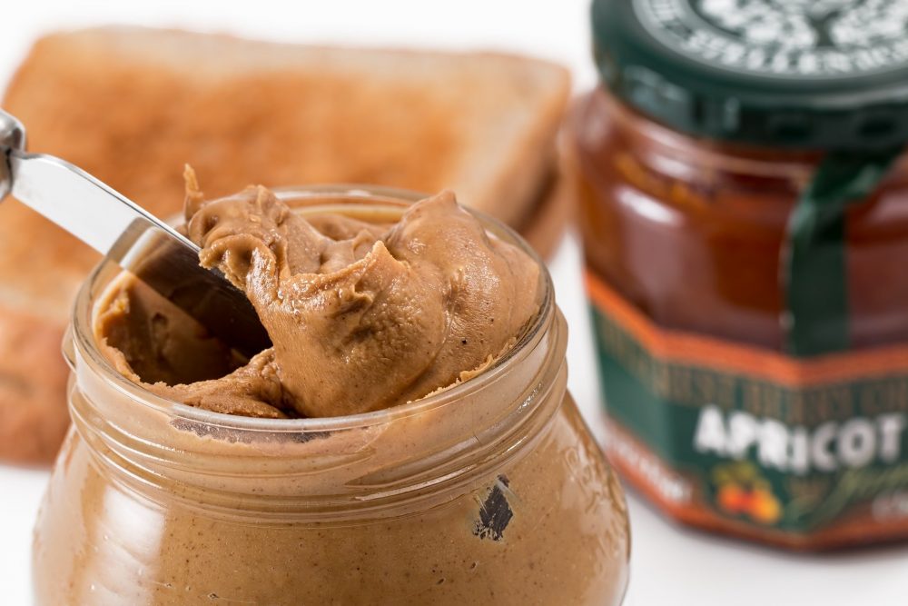 A knife in a peanut butter fat with toast and apricot jam in the background