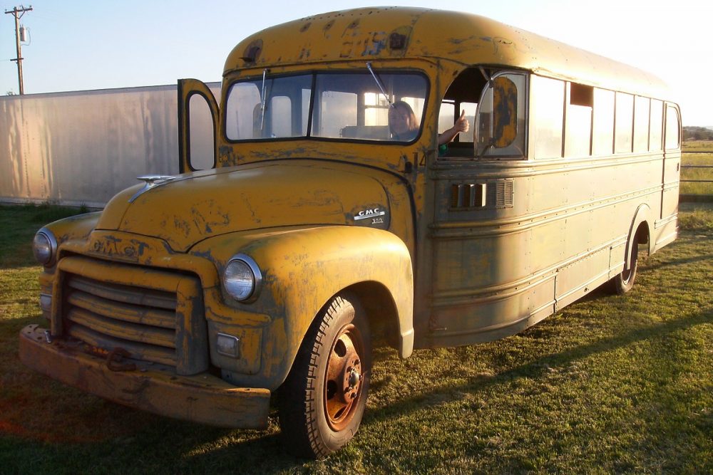 A woman gives a thumbs up from the front seat of a rusted 1955 school bus parked in grass