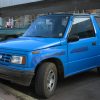 A bright blue 1991 Geo Tracker Convertible is parked in front of a shop