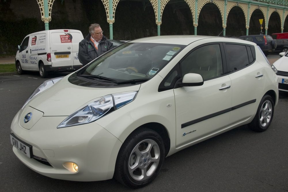 A white 2011 Nissan LEAF is parked with its lights on while a man talks to the driver