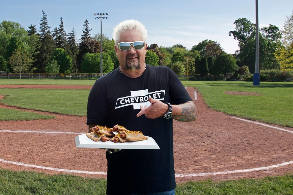 Guy Fieri standing at the Field of Dreams in Iowa and holding a plate of Apple Pie Hot Dogs