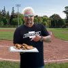 Guy Fieri standing at the Field of Dreams in Iowa and holding a plate of Apple Pie Hot Dogs