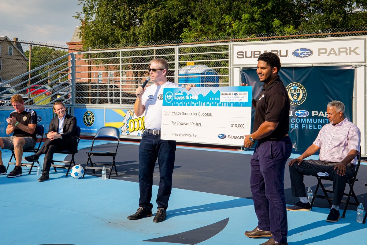 A Subaru employee presents a $10,000 check to a Philadelphia YMCA employee at Subaru Park Camden Mini-Pitch