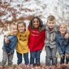 group of five children wearing coats and standing on a pile of autumn leaves with a tree in the background