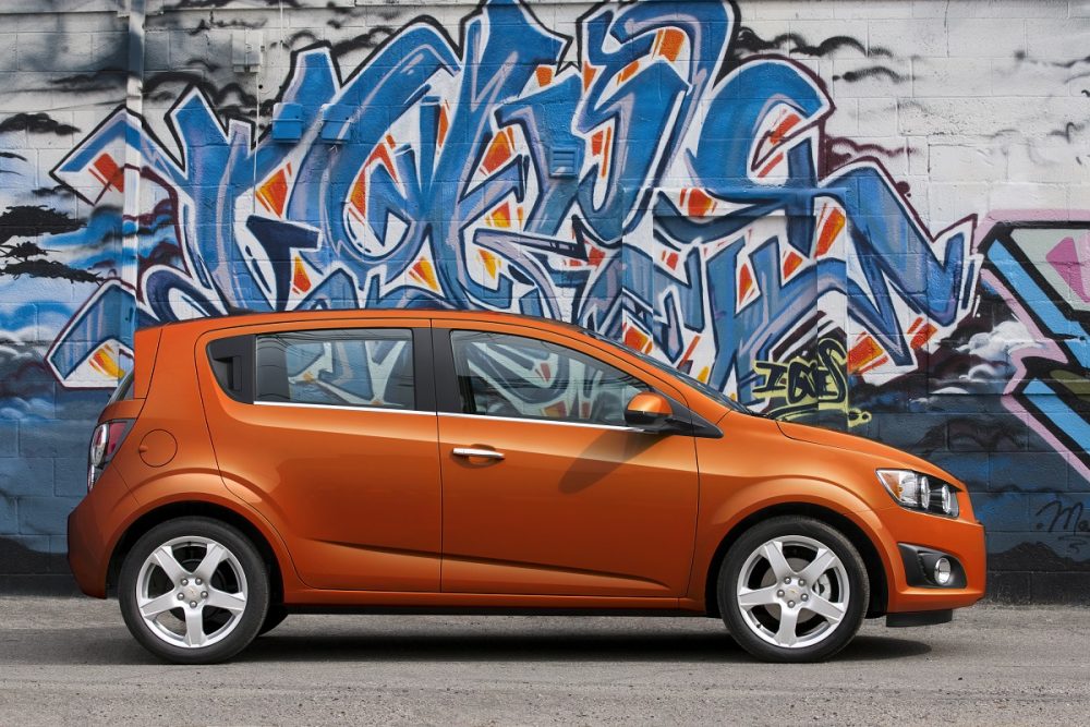 An orange 2012 Chevrolet Sonic Hatchback in front of a wall with graffiti