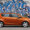 An orange 2012 Chevrolet Sonic Hatchback in front of a wall with graffiti