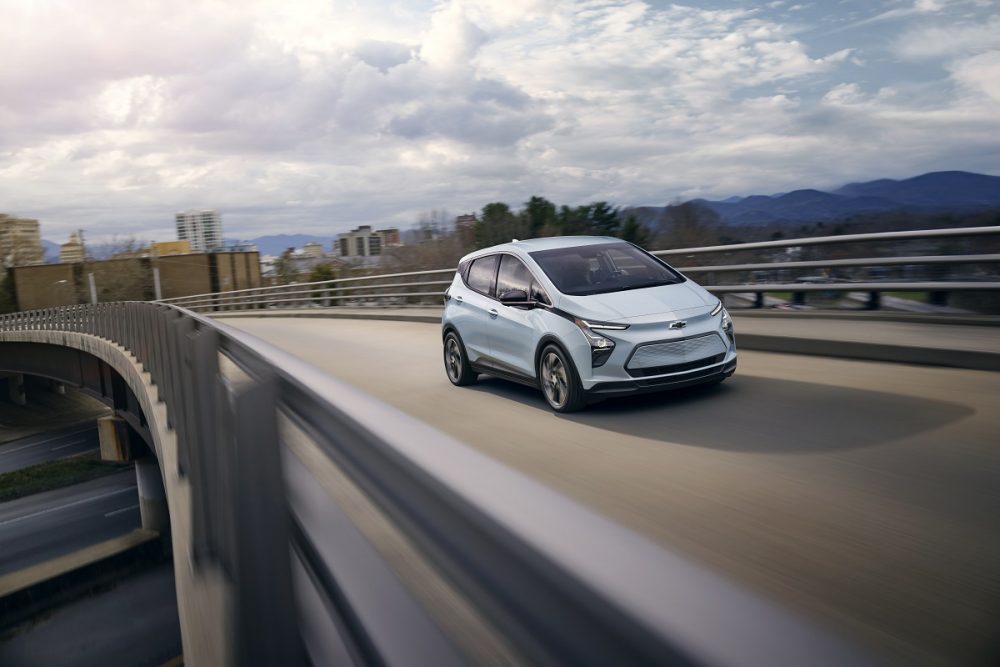 Front side view of 2023 Chevrolet Bolt EV driving down across bridge with city skyline in background