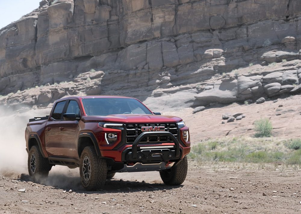 Front side view of red 2023 GMC Canyon AT4X kicking up dust cloud on gravel trail against rocky cliff backdrop