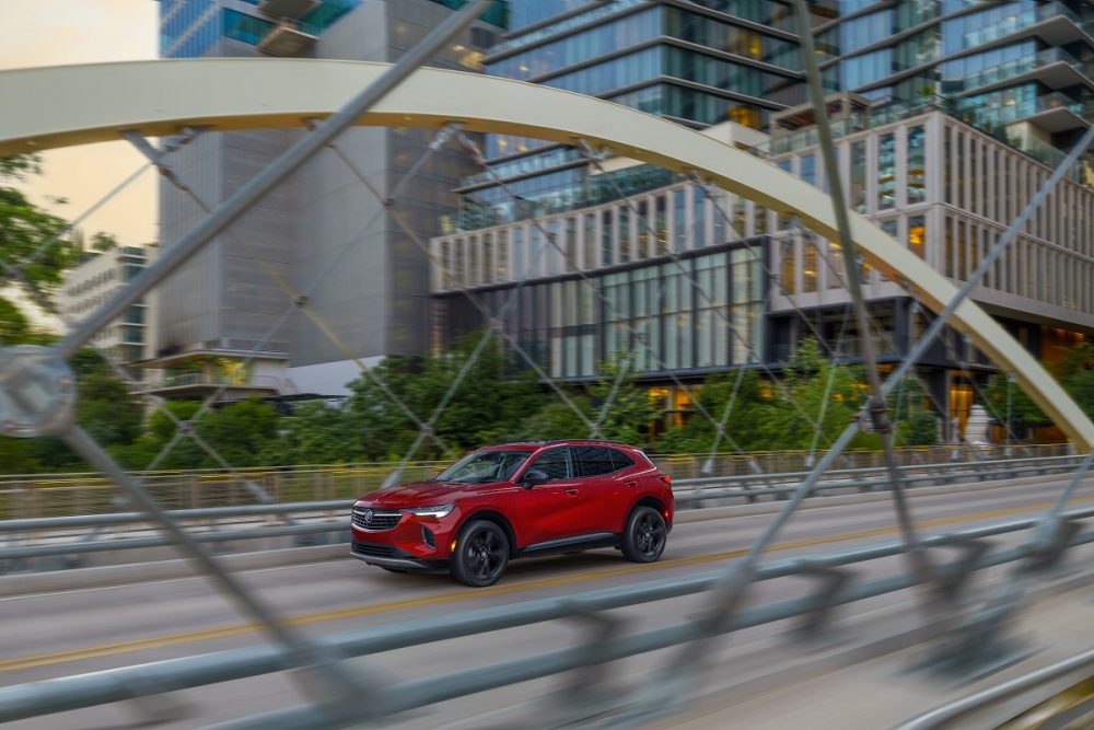 Front side view of red 2023 Buick Envision driving on city street with fence in foreground and buildings in background