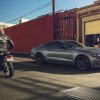 A man sitting astride a motorcycle looks over at a 2024 Ford Mustang GT parked on the street
