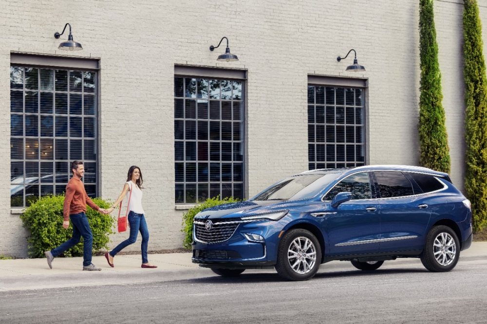 Front side view of blue 2024 Buick Enclave parked in front of building as two people approach from the left