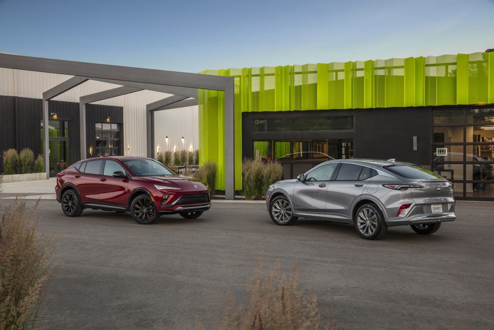 Two 2024 Buick Envista models are parked nose to nose in front of a building. A Buick Envista Sport Touring in Cinnabar Metallic is on the left, and a Buick Envista Avenir in Moonstone Gray Metallic is on the right.