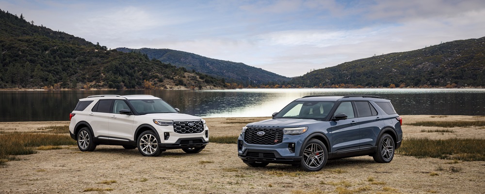 A 2025 Ford Explorer Platinum in Star White Metallic Tri-Coat and a 2025 Ford Explorer ST in Vapor Blue Metallic sit side by side on a lakefront