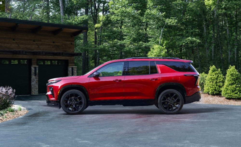 Driver’s side profile view of 2024 Chevrolet Traverse RS in Radiant Red Tintcoat parked in a driveway with a garage and woods in the background