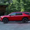 Driver’s side profile view of 2024 Chevrolet Traverse RS in Radiant Red Tintcoat parked in a driveway with a garage and woods in the background