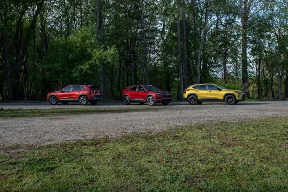 Side views of the 2024 Chevrolet Trax LT in Cayenne Orange Metallic, RS in Crimson Metallic, and ACTIV in Nitro Yellow Metallic parked on a gravel road in front of trees