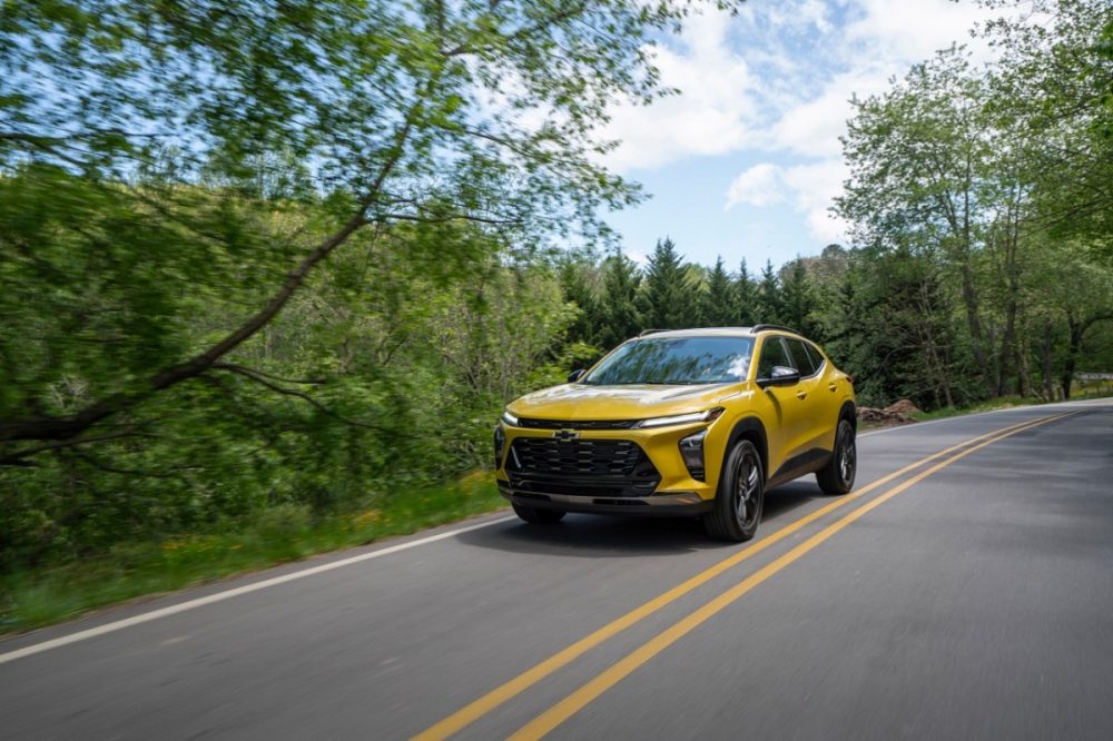 Front 3/4 view of 2024 Chevrolet Trax ACTIV in Nitro Yellow Metallic driving on a road lined with trees