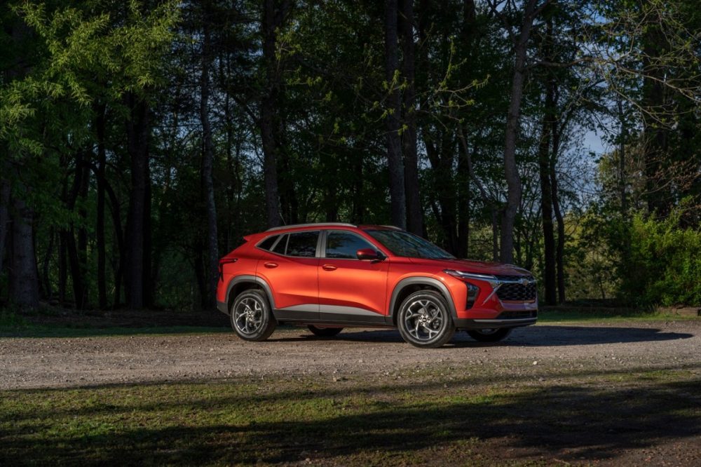 Passenger’s side 7/8 view of 2024 Chevrolet Trax LT in Cayenne Orange Metallic parked on a gravel road in front of trees