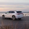 Rear 3/4 shot of a white 2025 Buick Enclave Avenir parked with tall grass in the foreground and a beach and water in the background