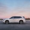 Side profile view of a white 2025 Buick Enclave Avenir parked on asphalt with grassy dunes and evening sky in the background