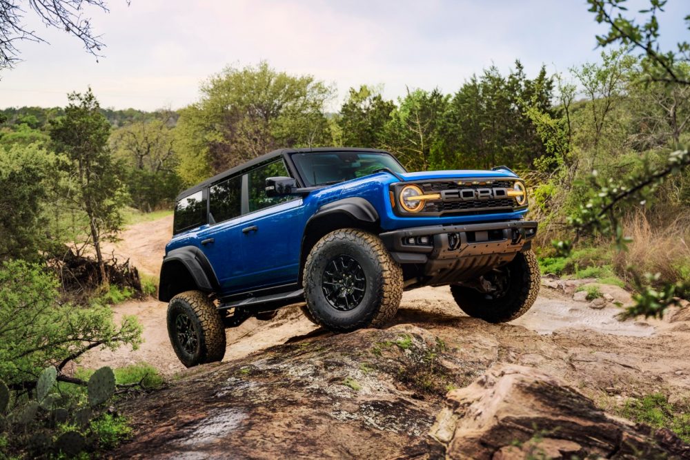 A 2024 Ford Bronco Raptor in Velocity Blue Metallic with the Black Appearance Package climbs over uneven terrain in a woodland