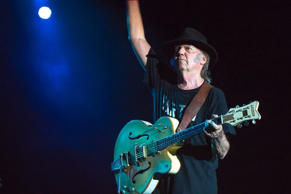 During a 2014 concert, Neil Young raises one hand while grasping the neck of his guitar with the other