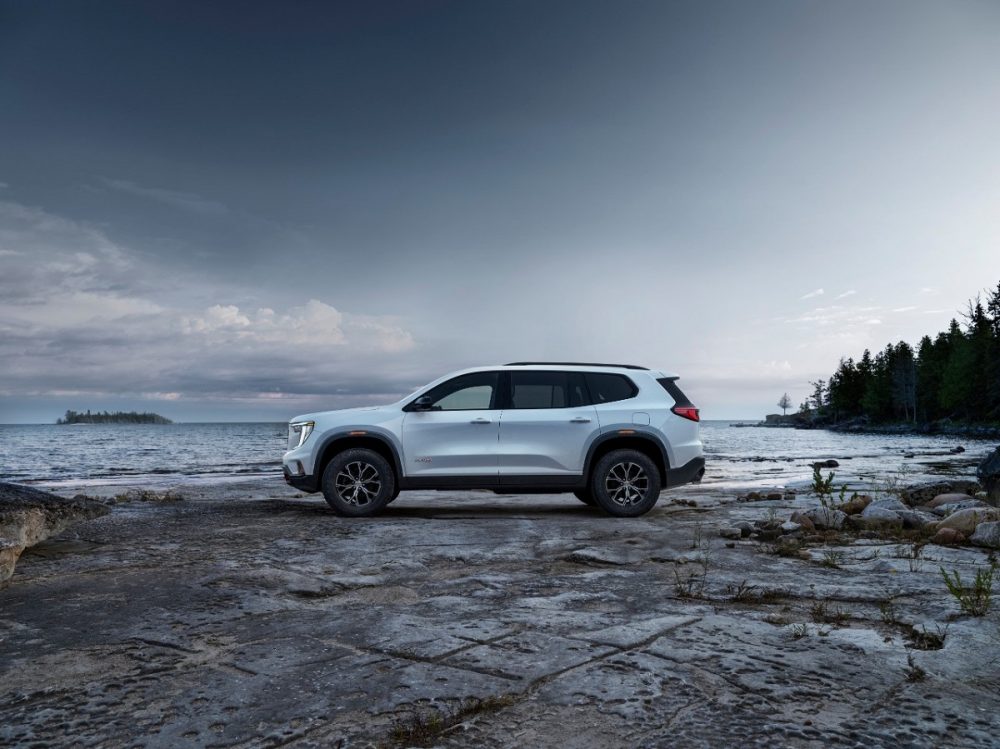 A full side profile view of the 2024 GMC Acadia AT4 parked on the rocky, wooded shores of a lake