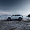 A full side profile view of the 2024 GMC Acadia AT4 parked on the rocky, wooded shores of a lake