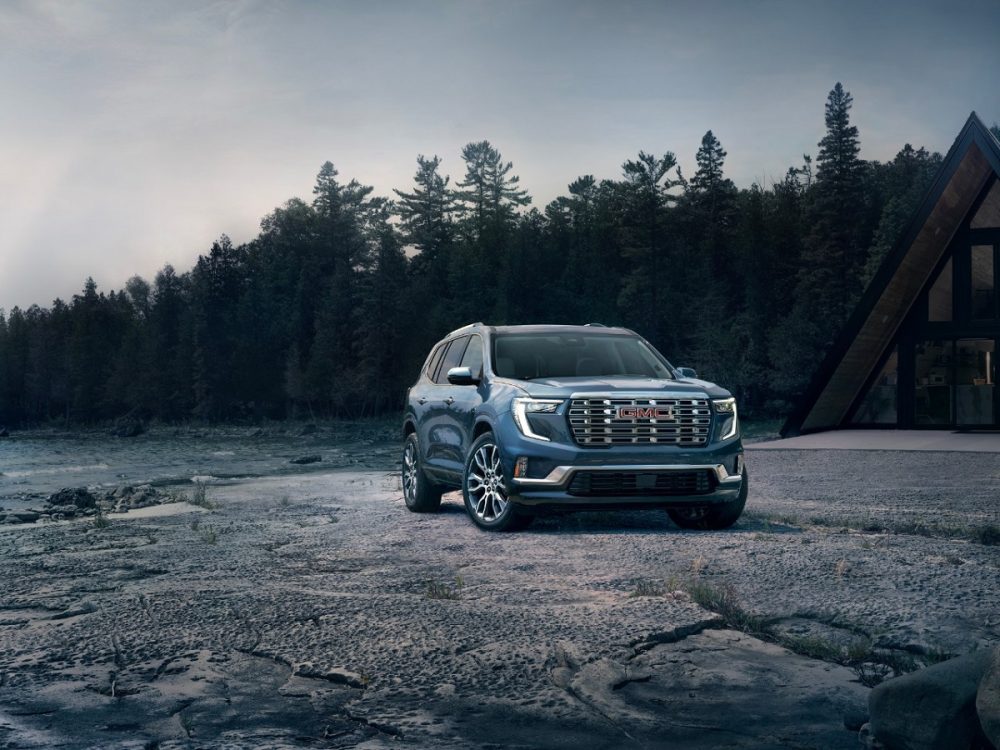 A front 3/4 view of the 2024 GMC Acadia Denali parked on the rocky, wooded shore of a lake