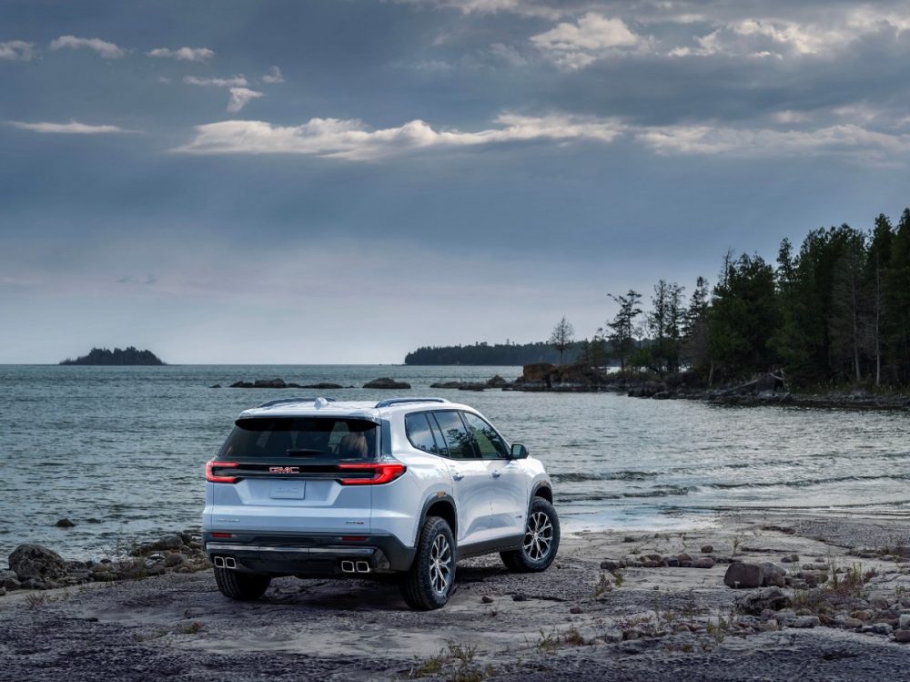 A rear 3/4 view of the 2024 GMC Acadia AT4 parked on the rocky, wooded shore of a lake