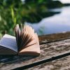 A breeze ruffles the pages of an open book lying on a dock next to a body of water