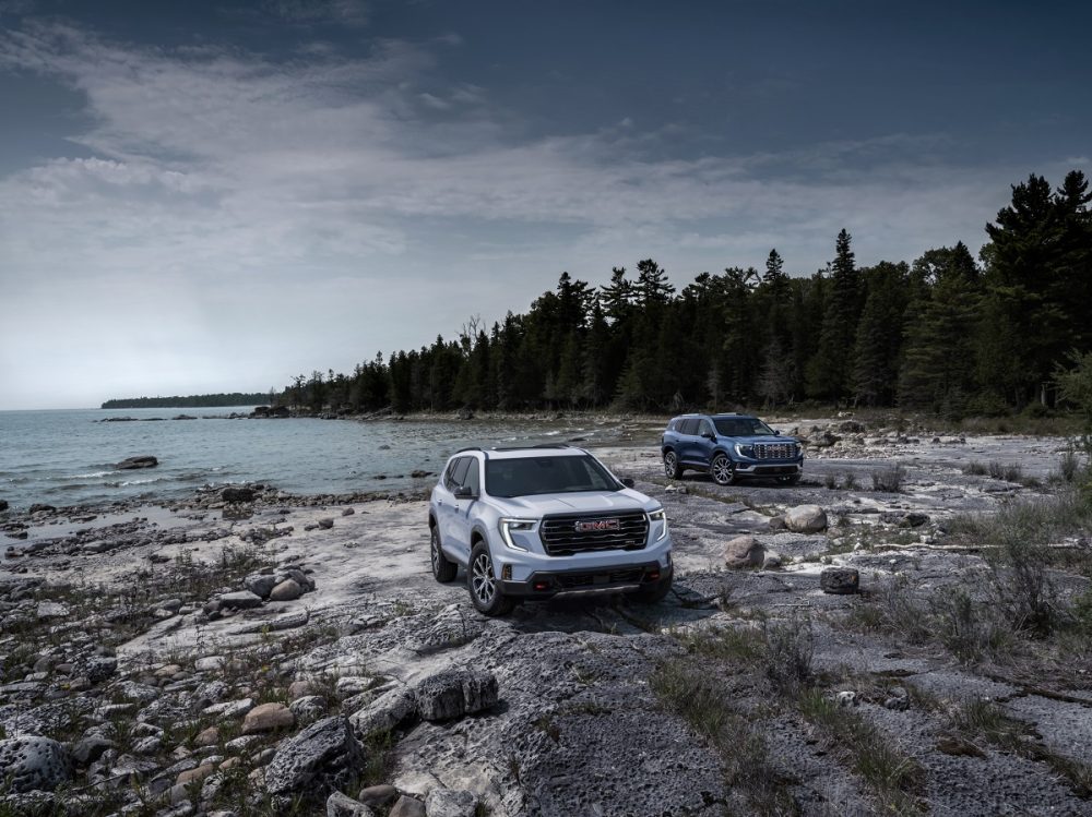 The 2024 GMC Acadia AT4 (front) and 2024 GMC Acadia Denali (back) sit on a rocky lakeshore with water and tall trees in the background