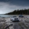 The 2024 GMC Acadia AT4 (front) and 2024 GMC Acadia Denali (back) sit on a rocky lakeshore with water and tall trees in the background