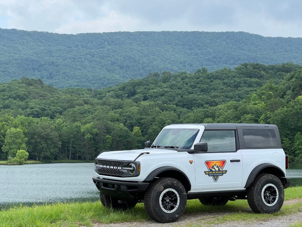 A Ford Bronco with the Bronco Off-Roadeo logo on the driver's-side door sits on the banks of a river near the Great Smoky Mountains