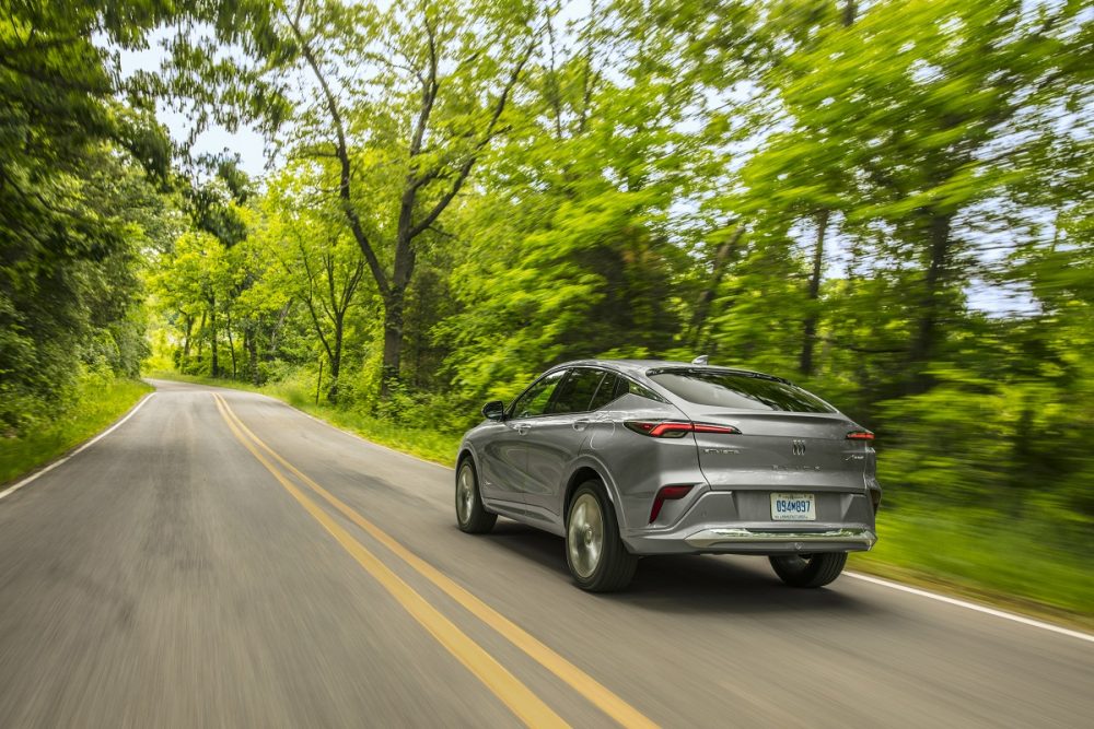 Rear 3/4 view of gray 2024 Buick Envista Avenir driving on tree-lined road