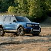 A blue 2025 Ford Bronco Sport drives over uneven terrain near a woodland.