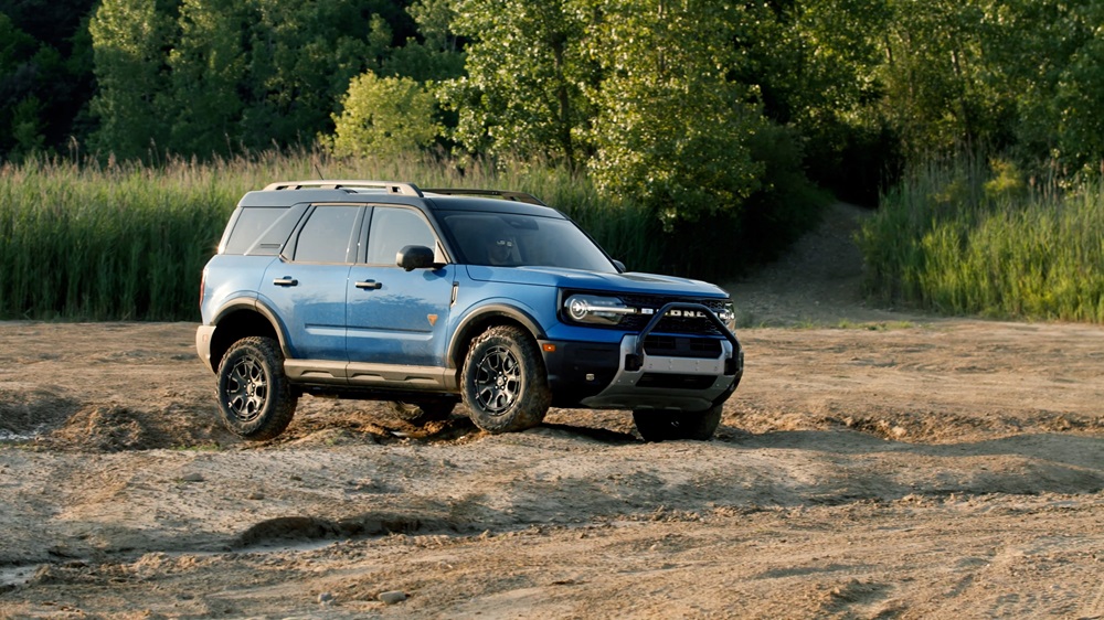 A blue 2025 Ford Bronco Sport drives over uneven terrain near a woodland.