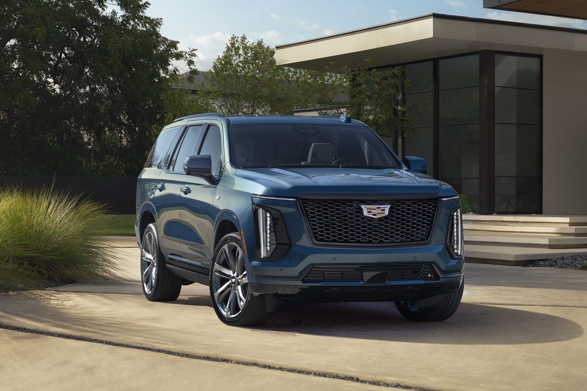 Front ¾ view of the 2025 Cadillac Escalade Sport Platinum in Deep Sea Metallic with a house and greenery in the background.