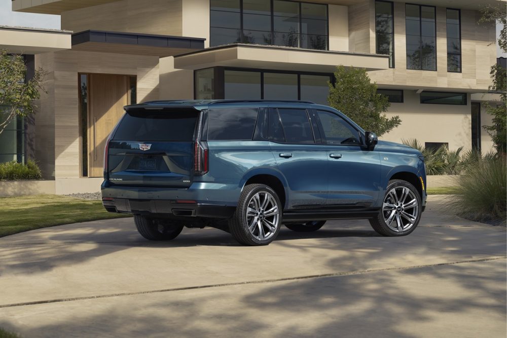 Rear ¾ view of the 2025 Cadillac Escalade Sport Platinum in Deep Sea Metallic with a house and greenery in the background.