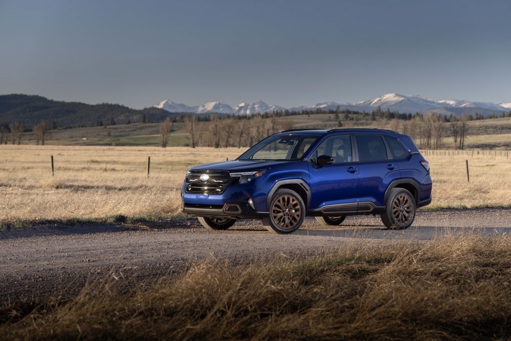 A blue 2025 Subaru Forester parked on a gravel road with fields and mountains in the background