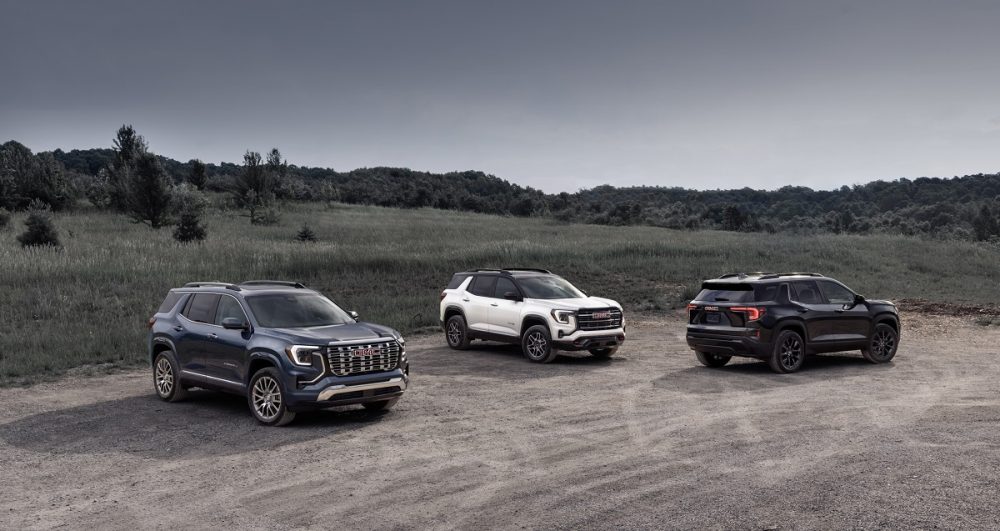 The 2026 GMC Terrain Denali, 2026 Terrain AT4, and 2025 Terrain Elevation are parked next to each other on a gravel lot with a field and trees in the background